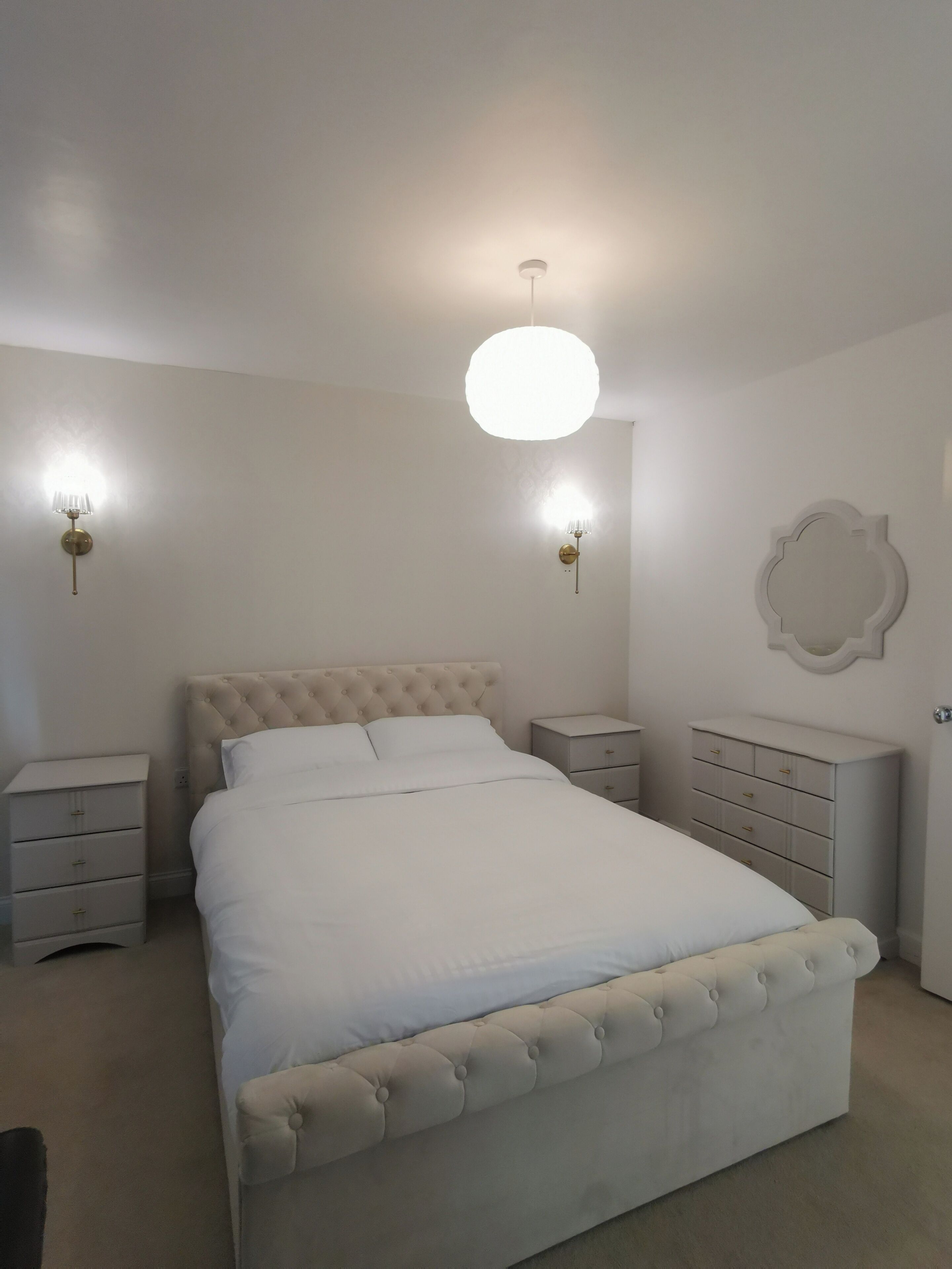 A bedroom with a large bed, two bedside tables, a wall-mounted light fixture, and a decorative mirror.