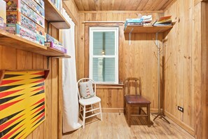 Una habitación pequeña con paredes de madera, una ventana con contraventanas, una silla blanca, un banco de madera y estanterías con libros y juegos.
