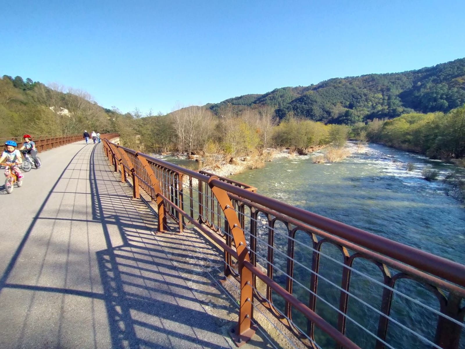A pedestrian bridge with a metal railing, a river below, and a hilly landscape in the background.