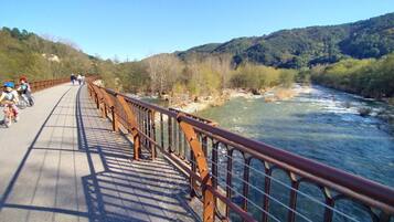 A pedestrian bridge with a metal railing, a river below, and a hilly landscape in the background.
