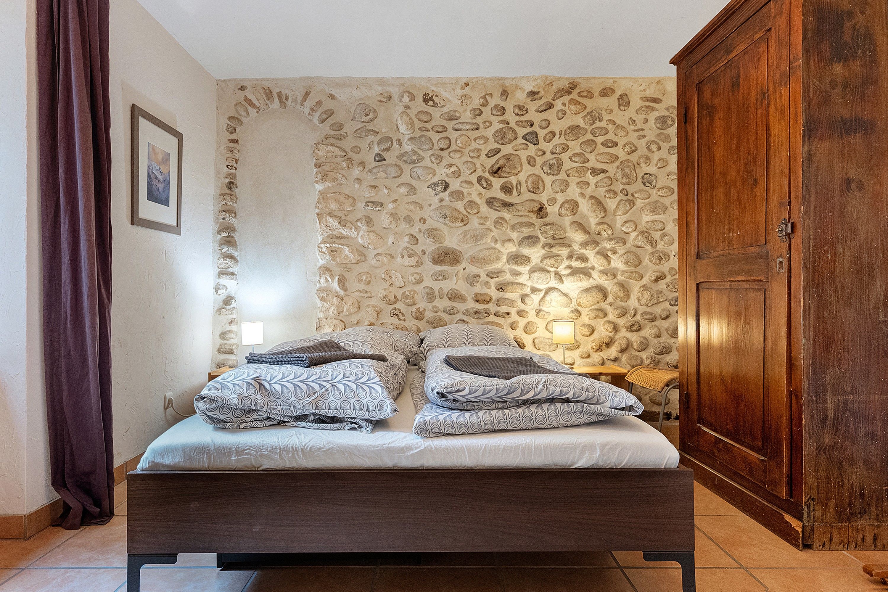 Ein Schlafzimmer mit Steinsandsteinwand, einem Holzschrank und einem Bett mit gemusterten Bettbezügen.