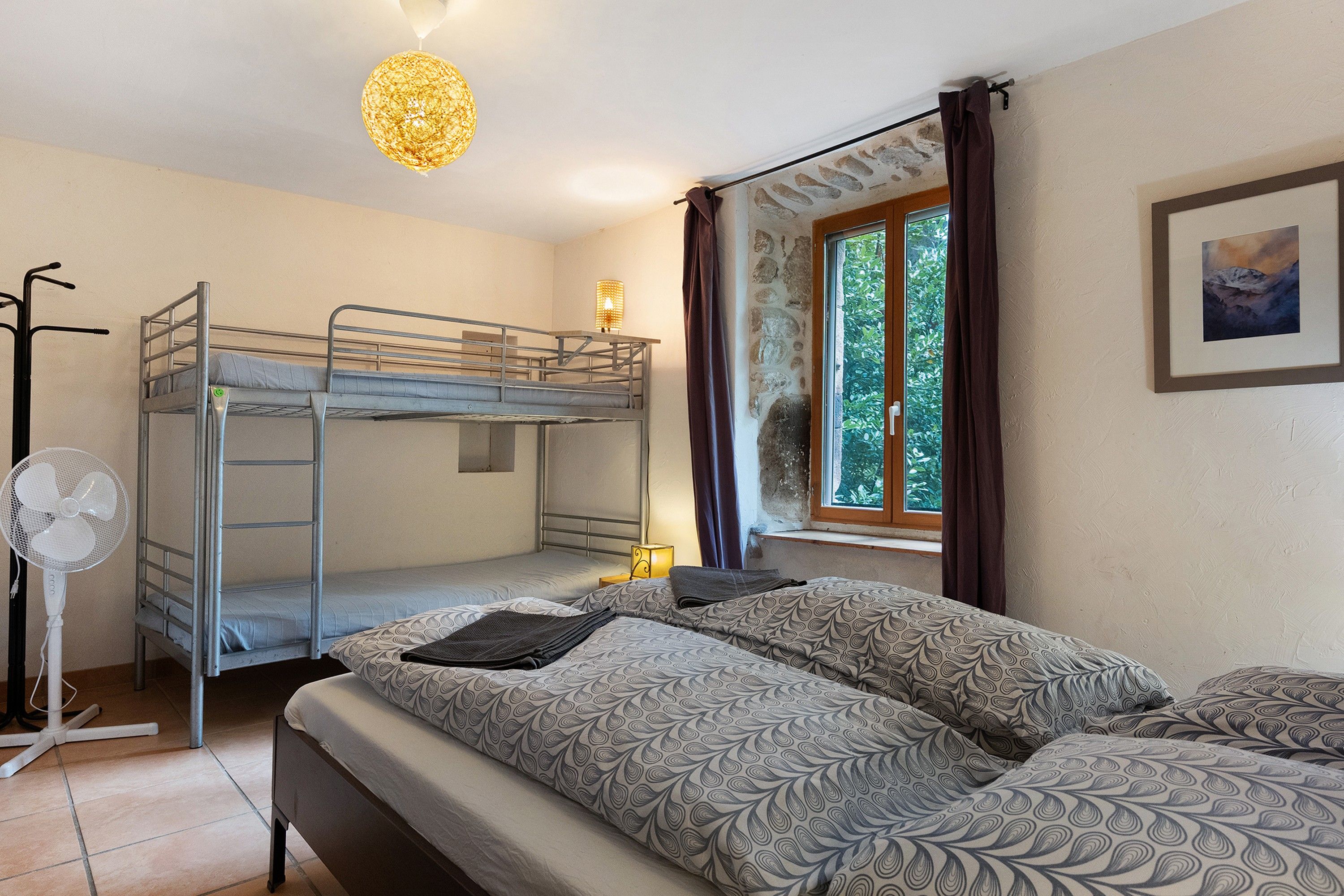 A room with bunk beds, a standing fan, a table lamp, a window with curtains, and a framed picture on the wall.