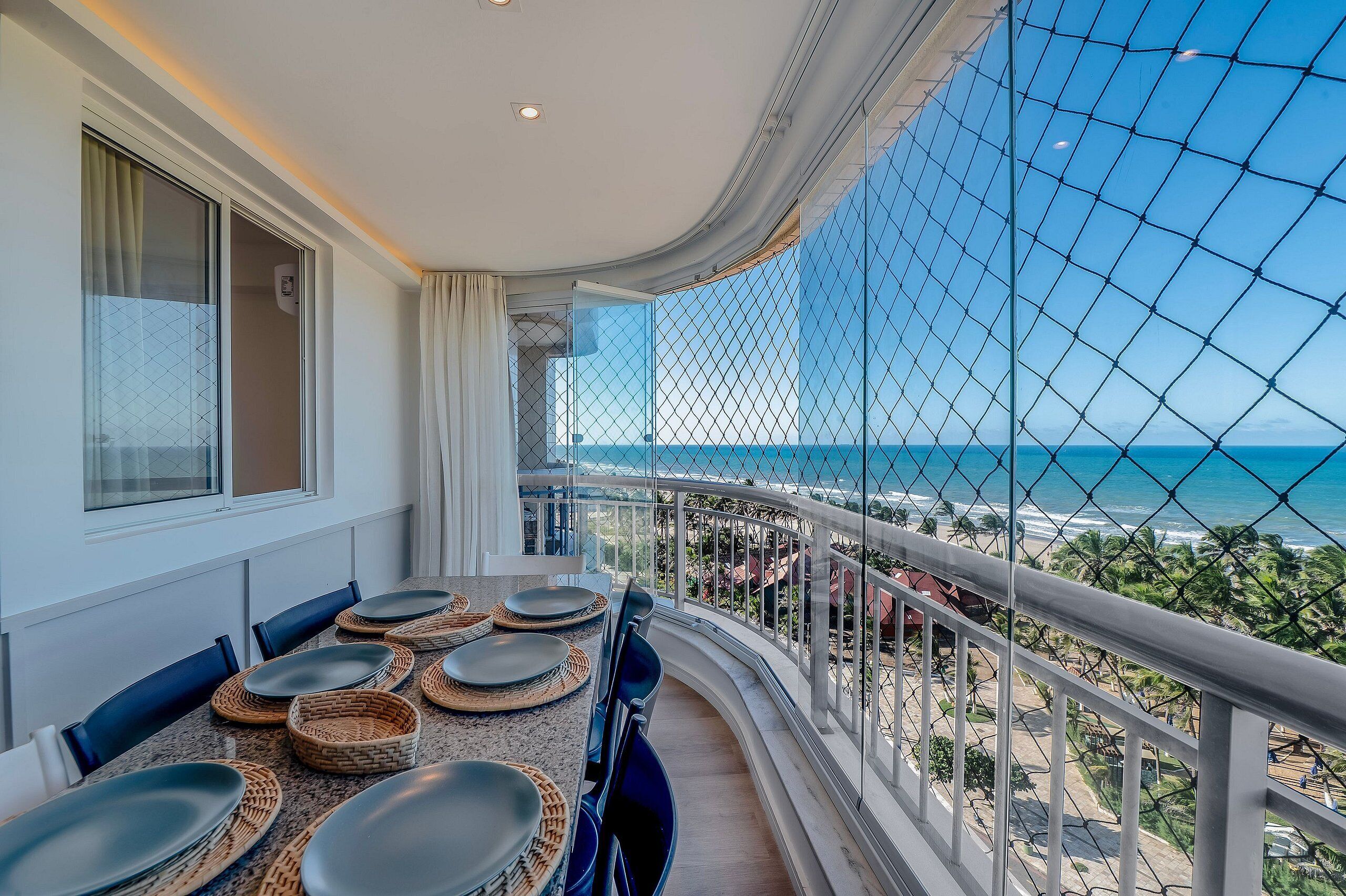 Varanda com vista para a praia, mesa de jantar com pratos e cadeiras, e guarda-corpo de vidro.