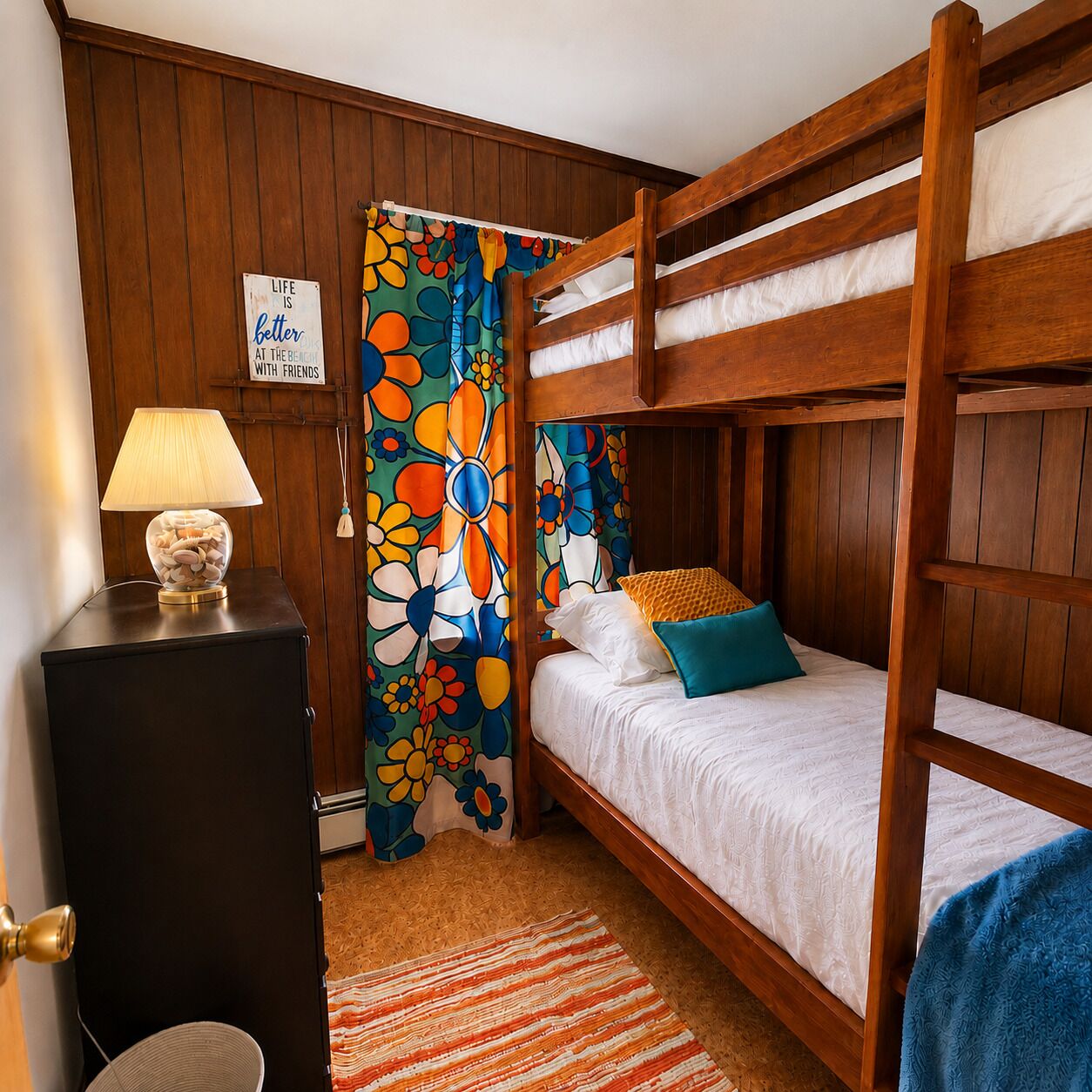 A bunk bed room with a wooden headboard, a bedside table with a lamp, a colorful floral curtain, and a sign on the wall.