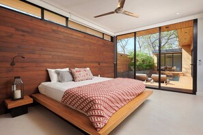 Un dormitorio moderno con cabecera de madera, una cama grande, una mesita de noche con una linterna y una puerta corrediza de cristal que da a un espacio exterior.