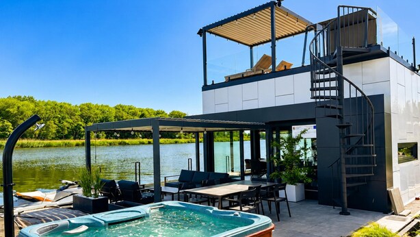 A modern outdoor hot tub area with a wooden deck, seating, and a view of a lake. - Boat House for 7 With Jacuzzi (Dziwnów)