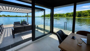 A modern outdoor deck with a view of a lake, furnished with a table and chairs, and a covered seating area. - Boat House for 7 With Jacuzzi (Dziwnów)