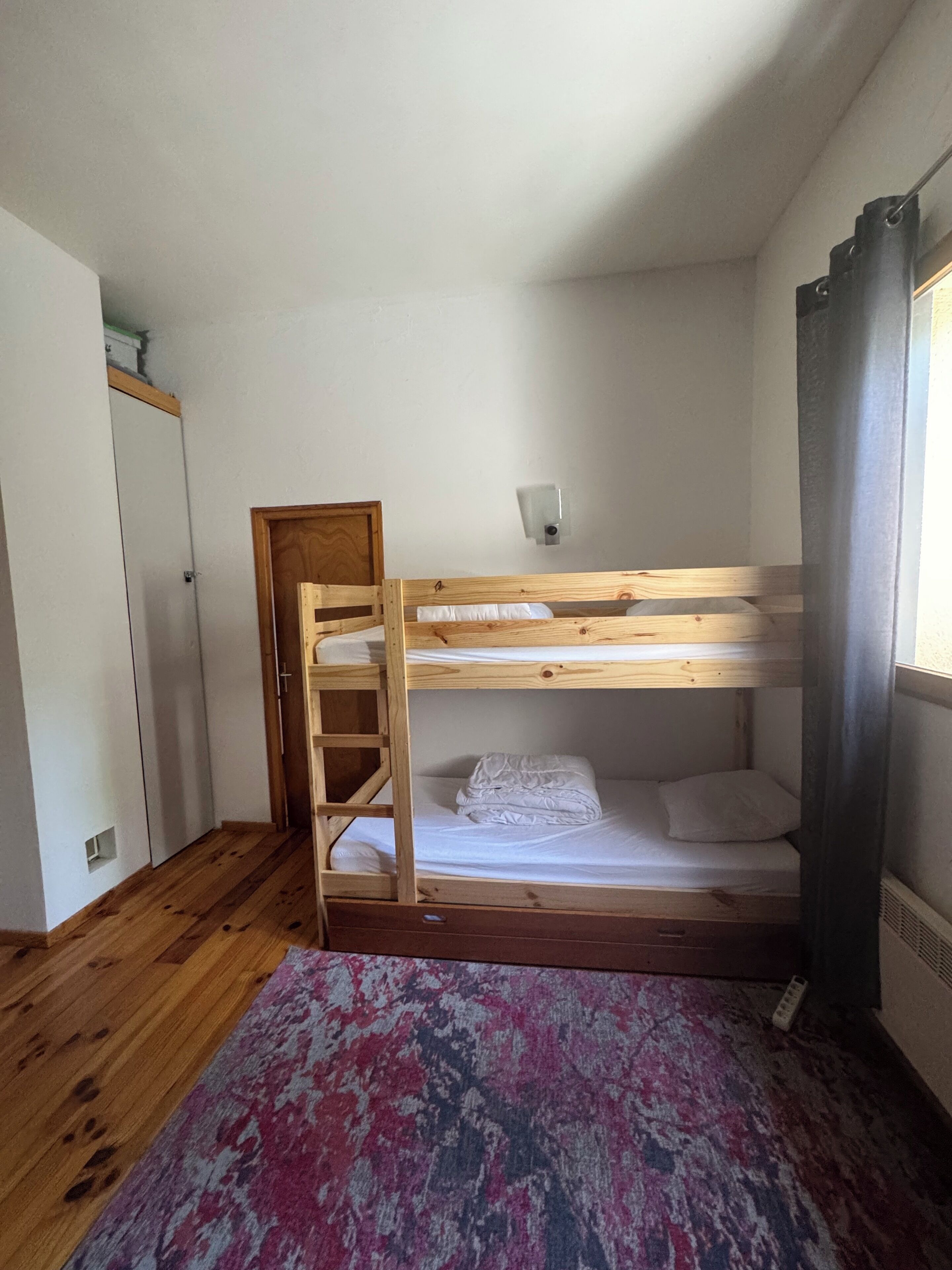 Camera con letto a castello, una scala in legno, una piccola finestra con tende e un tappeto a motivi sul pavimento.