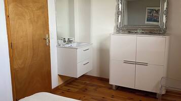 Une chambre avec une porte en bois, une table de chevet blanche, une commode blanche avec un miroir et un lit recouvert d’une couette blanche.