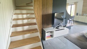 Eine Treppe mit Holzbodenstufen führt zu einer Zwischenebene, auf einer weißen Unterhaltungsanlage befindet sich ein Flachbildfernseher, und das moderne Interieur zeichnet sich durch eine hohe Decke und einen Ausblick ins Freie aus.