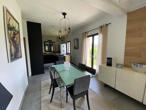 A modern dining area with a glass table, dark chairs, a kitchen in the background, and a view of greenery through the window. - Contemporary home with a view of the mountains (Pont de Montvert - Sud Mont Lozère)