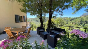 Eine Terrrasse im Freien mit einem Holztisch und -stühlen, einer schwarzen Rattan-Couch und Blick auf einen bewaldeten Hang.