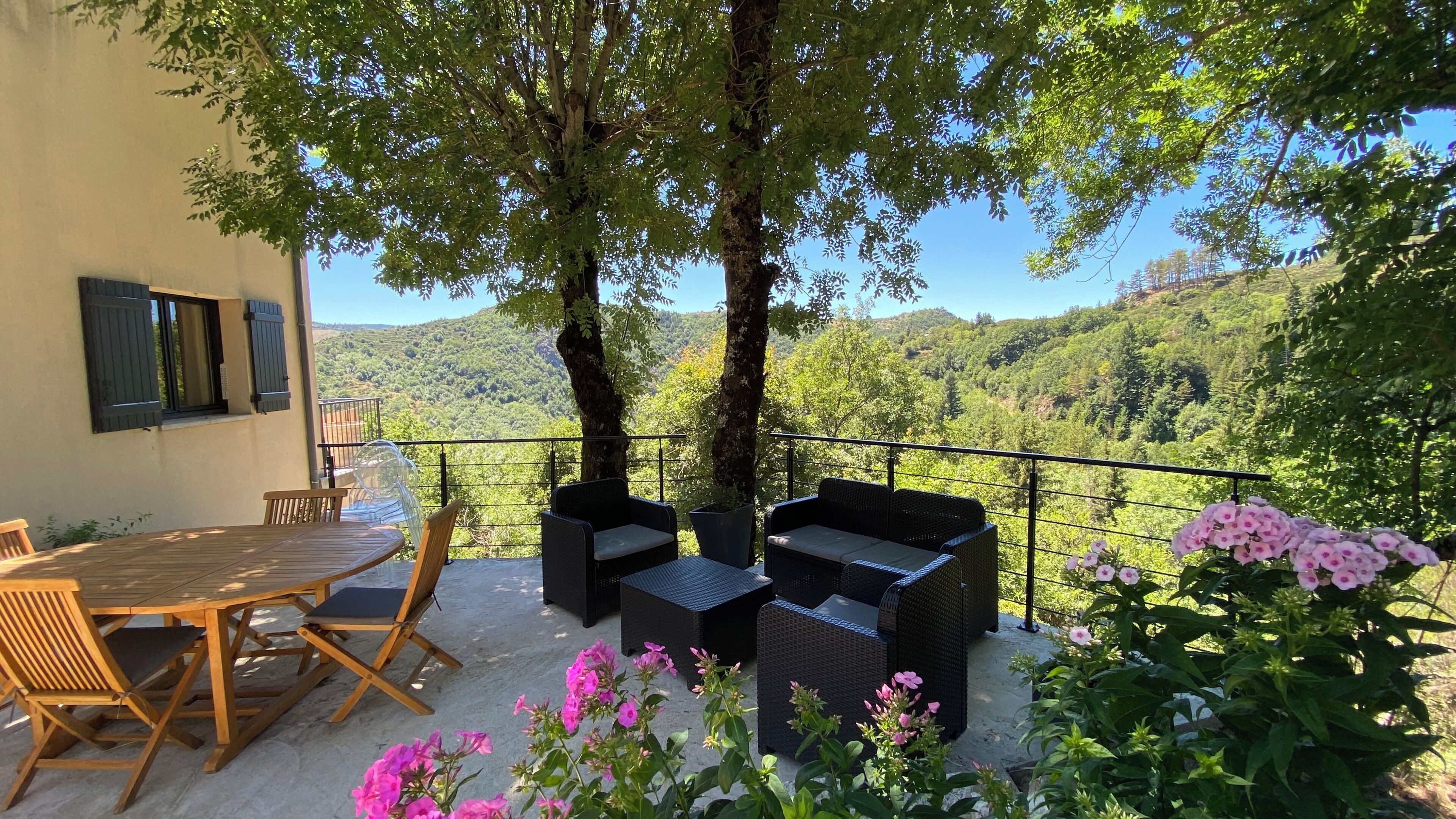 A terrace with a table and chairs, a wicker sofa, and a view of a forested hillside.