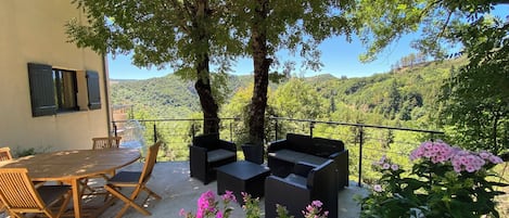 Una terraza con mesa y sillas, un sofá de mimbre y vistas a una ladera boscosa.