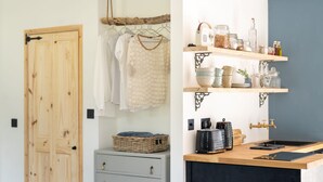 Una habitación con puerta de madera, un aparador gris con cajones, una cesta de mimbre y una zona de cocina con estantes, frascos y una cafetera.
