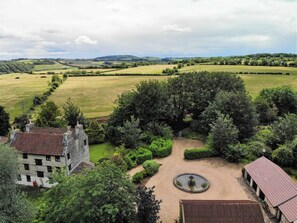 A rural landscape with a stone building, a circular garden with a fountain, and rolling green fields. - Country Cottage Bluebell (Keynsham)
