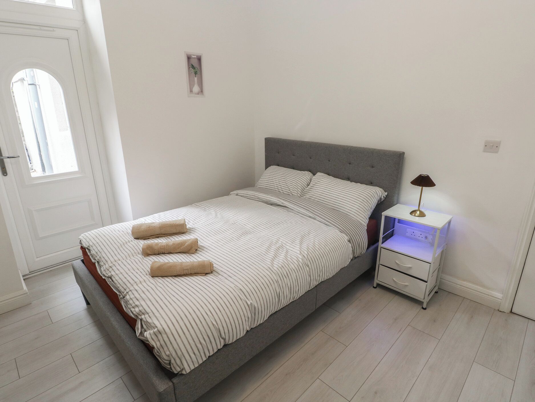 A single bed with striped bedding, a bedside table with a lamp, and a small plant on the wall.