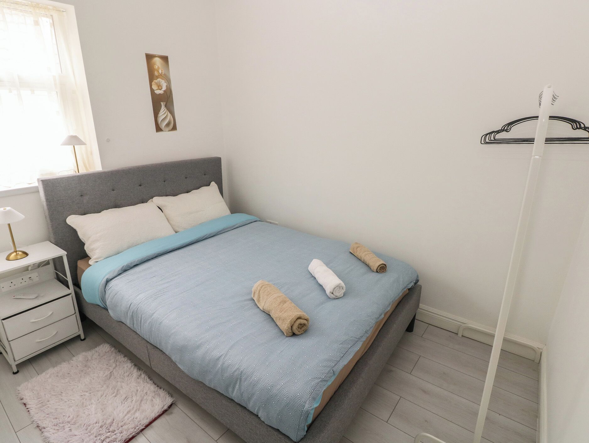 Una cama bien hecha con una colcha azul estampada, dos toallas enrolladas, una mesita de noche con una lámpara y una obra de arte colgada en la pared.