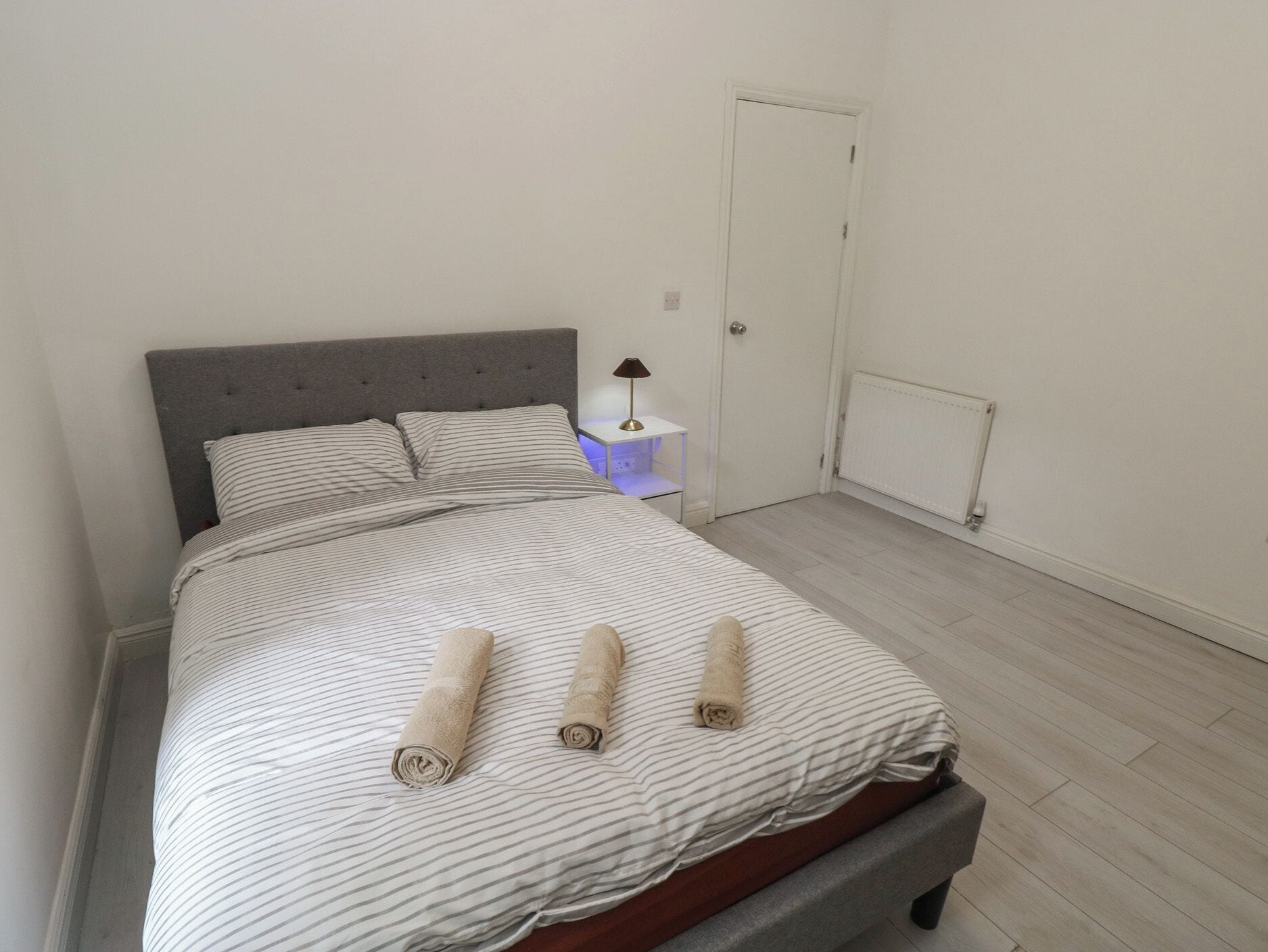 Een net opgemaakt bed met gestreepte lakens en opgerolde handdoeken, een nachtkastje met een lamp, en een witte deur in een minimalistische kamer.