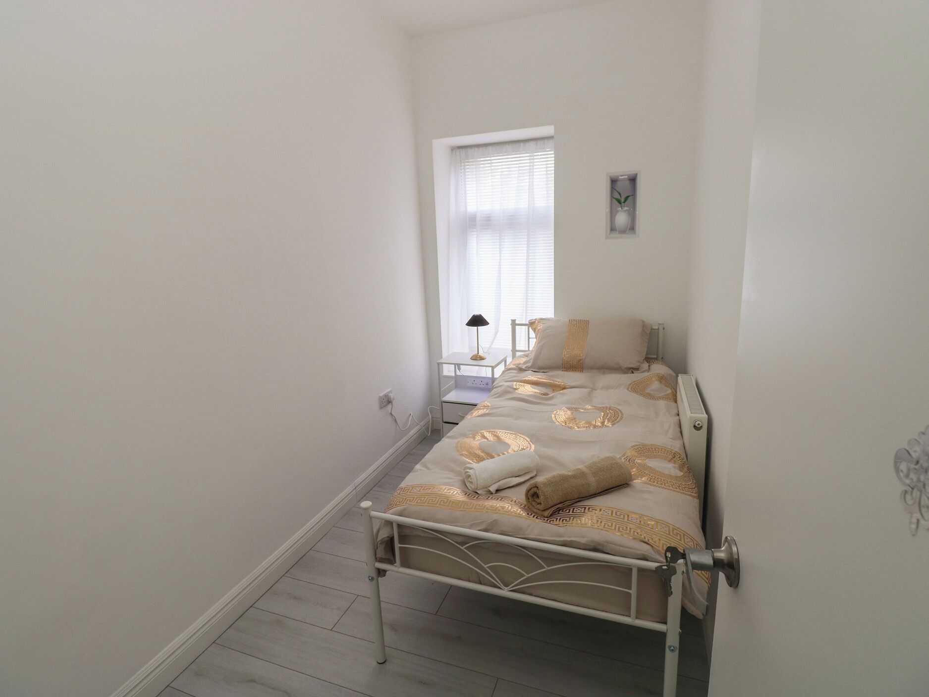 A narrow room with a single bed, a small bedside table, a window with blinds, and a wall-mounted light fixture.