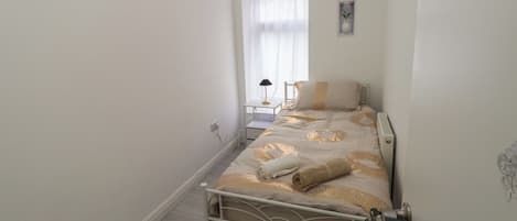 A narrow room with a single bed, a small bedside table, a window with blinds, and a wall-mounted light fixture.