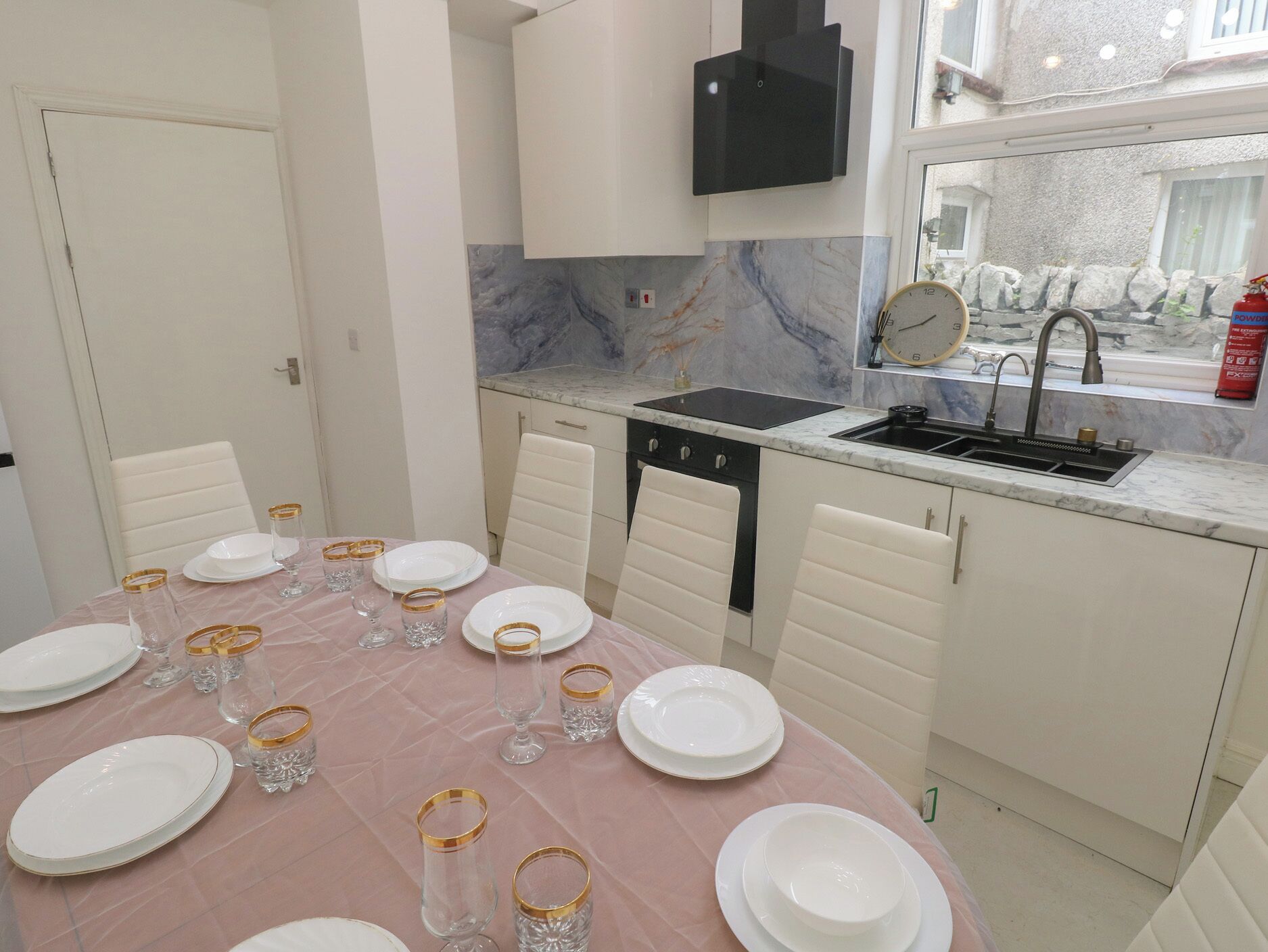A dining area with a round table set for six, white chairs, a black range hood, a marble backsplash, and a window with a view of the outside.
