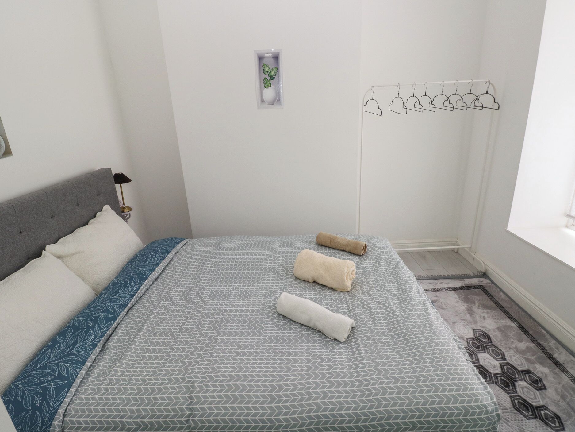 Un lit bien fait, recouvert d’une couvre-lit à motifs, deux serviettes enroulées, une lampe de chevet, une petite plante en pot et un étendoir fixé au mur.
