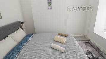 A neatly made bed with a patterned bedspread, two rolled towels, a bedside lamp, a small potted plant, and a wall-mounted drying rack.