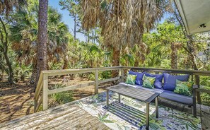 A wooden deck with a bench and table, surrounded by tropical vegetation. - Charming 3 Bedroom Cottage, Guest Cards + Cart! (Fripp Island)