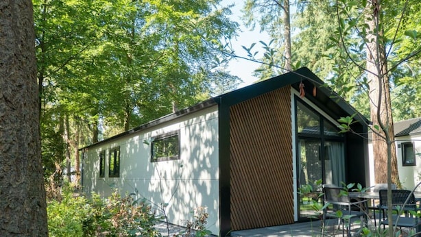A modern cabin with a large sliding glass door, surrounded by trees and a patio with outdoor furniture.