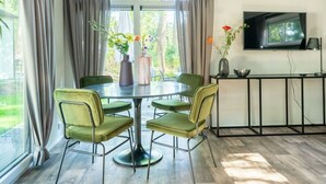 A dining area with a round table, green chairs, a TV on the wall, and a window with curtains.