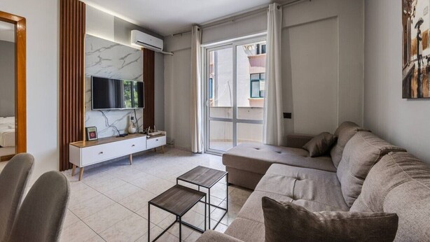 A modern living room with a sectional sofa, a TV mounted on a marble wall, a small table, and a large window with curtains. - Relax by the Sea | 1BR Apartment by PikHost (Durrës)