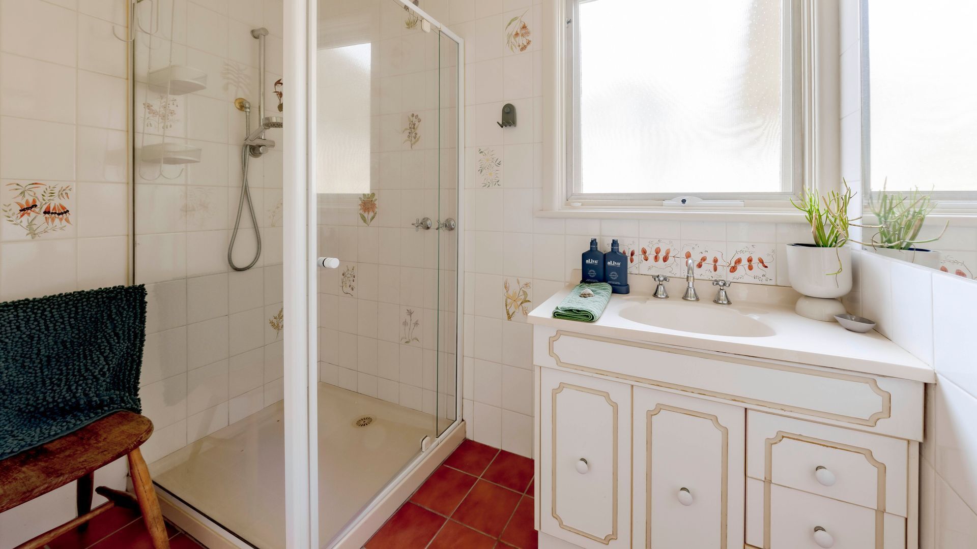 A bathroom with a glass shower enclosure, a sink with two faucets, and a window with plants.