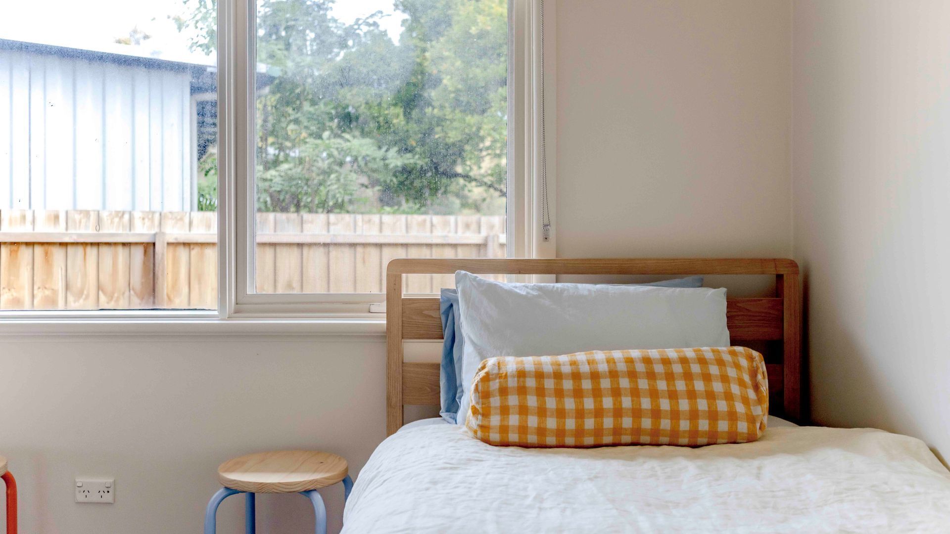A bedroom with a wooden bed, a small stool, a window with a view of a wooden fence and trees, and a white wall.
