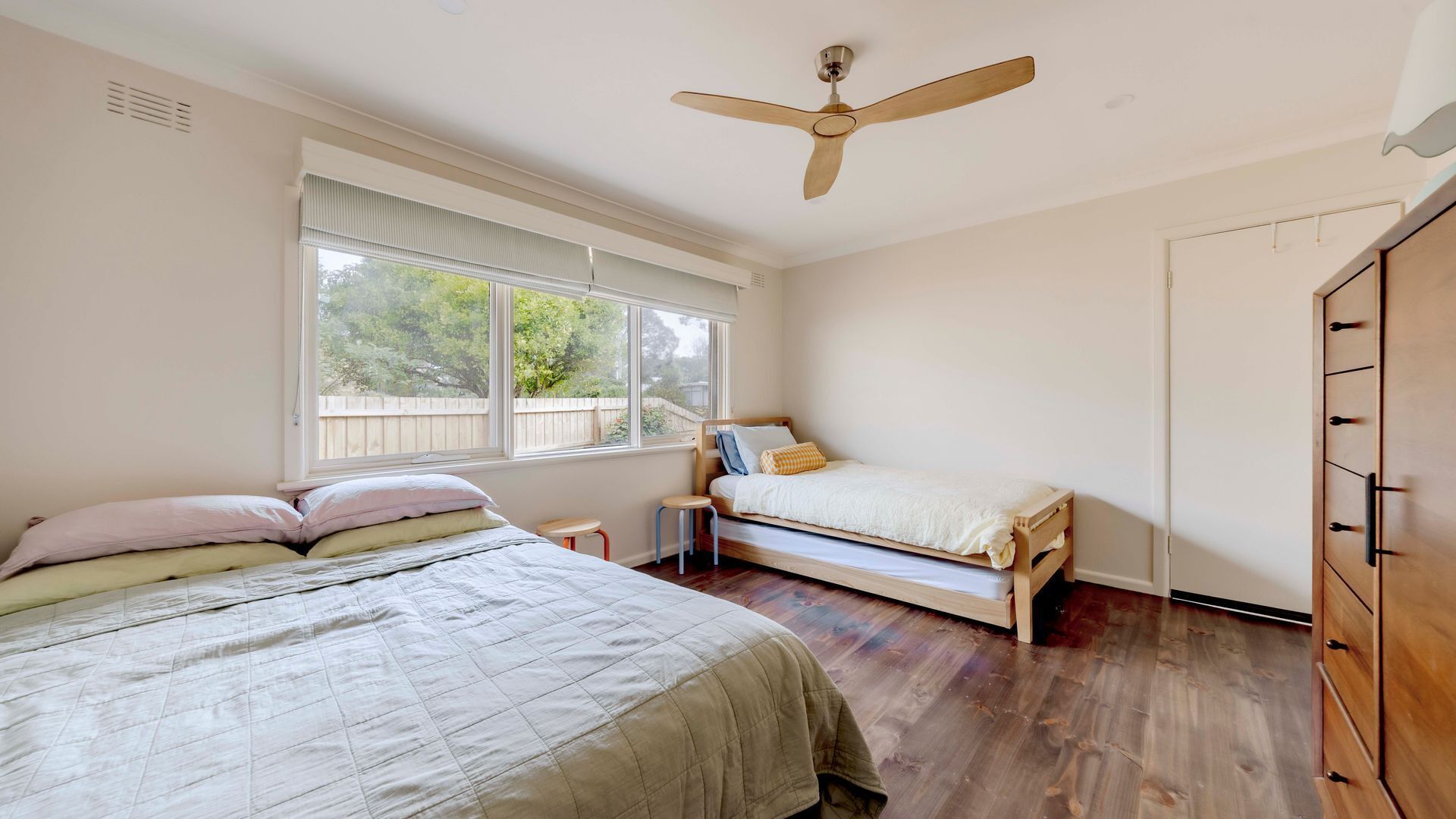 A bedroom with two beds, a ceiling fan, a window with a view, and a wooden wardrobe.