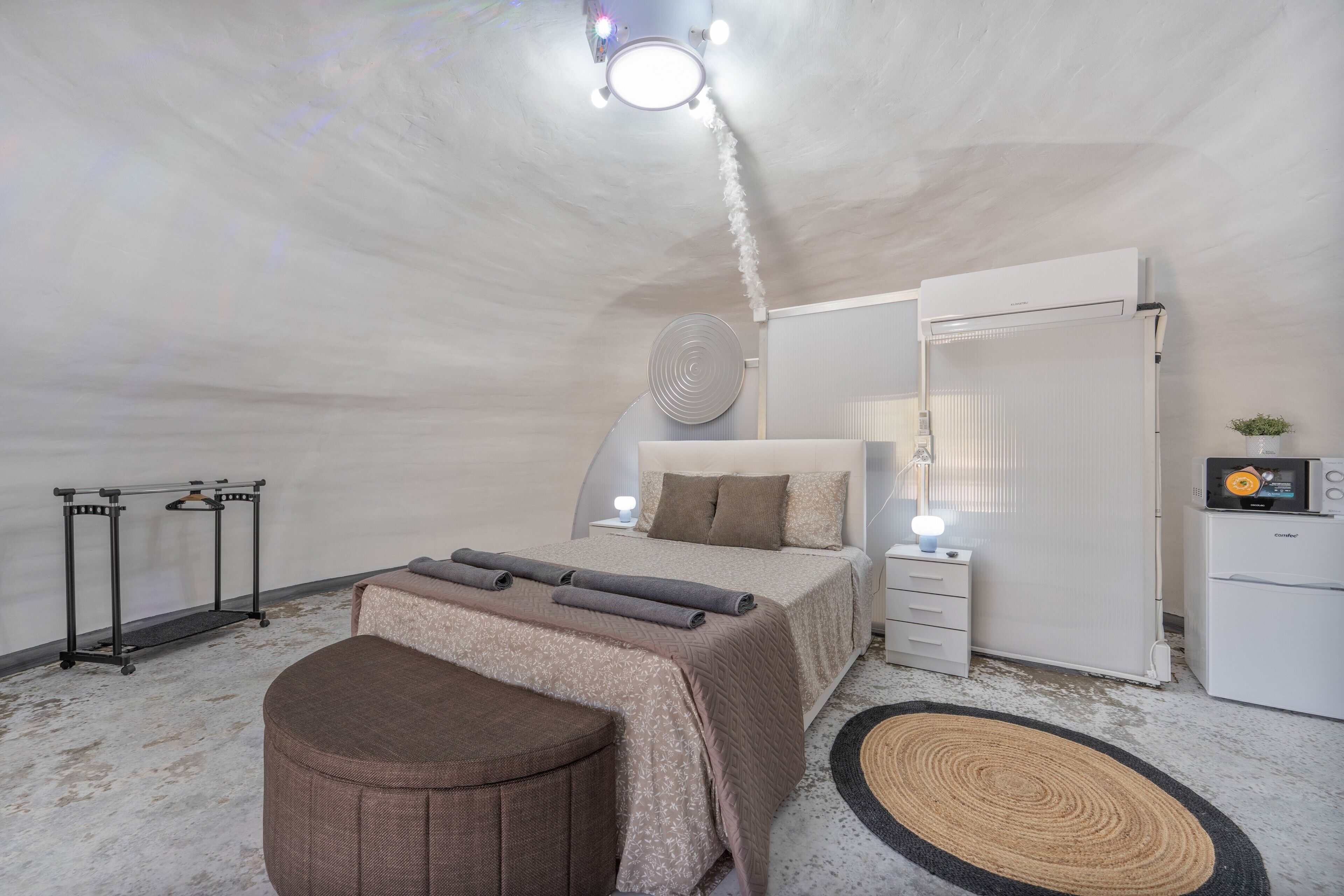 Un dormitorio con cama, mesitas de noche, armario, un pequeño otoman redondo, un radiador y una lámpara en el techo.