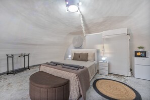 A bedroom with a bed, bedside tables, a wardrobe, a small round ottoman, a radiator, and a ceiling light fixture.