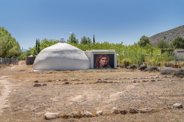 A dome-shaped structure with a mounted image on its side, surrounded by a dry landscape with sparse vegetation and a hill in the background.