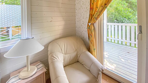 Une pièce d’angle avec un fauteuil confortable, une lampe et une fenêtre donnant sur une terrasse et de la verdure.