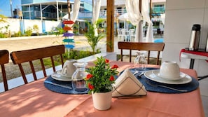 An outdoor dining area with a table set for two, a potted plant, and a view of a building with a blue and white facade.