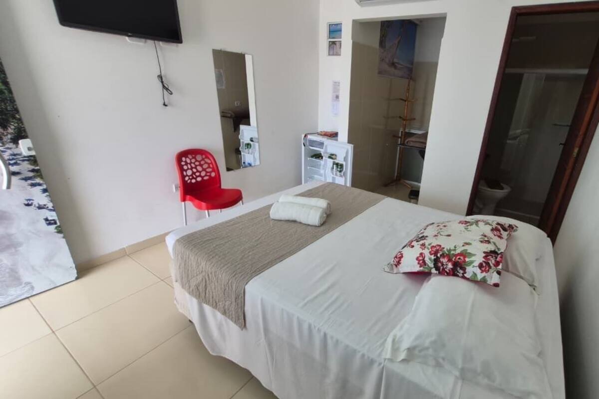 Un dormitorio con una cama, una silla roja, un armario blanco, un espejo y un televisor en la pared.
