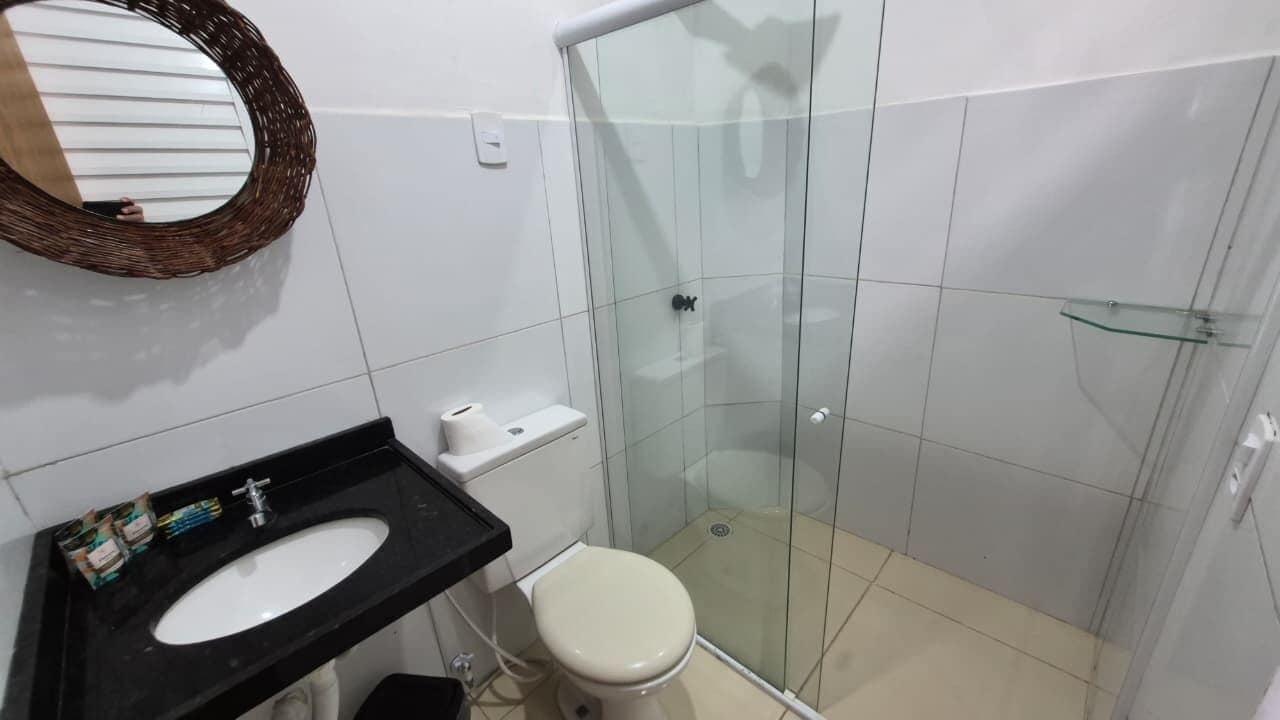 Un baño con lavabo negro, inodoro blanco, cabina de ducha con mampara de vidrio y espejo con marco de mimbre.