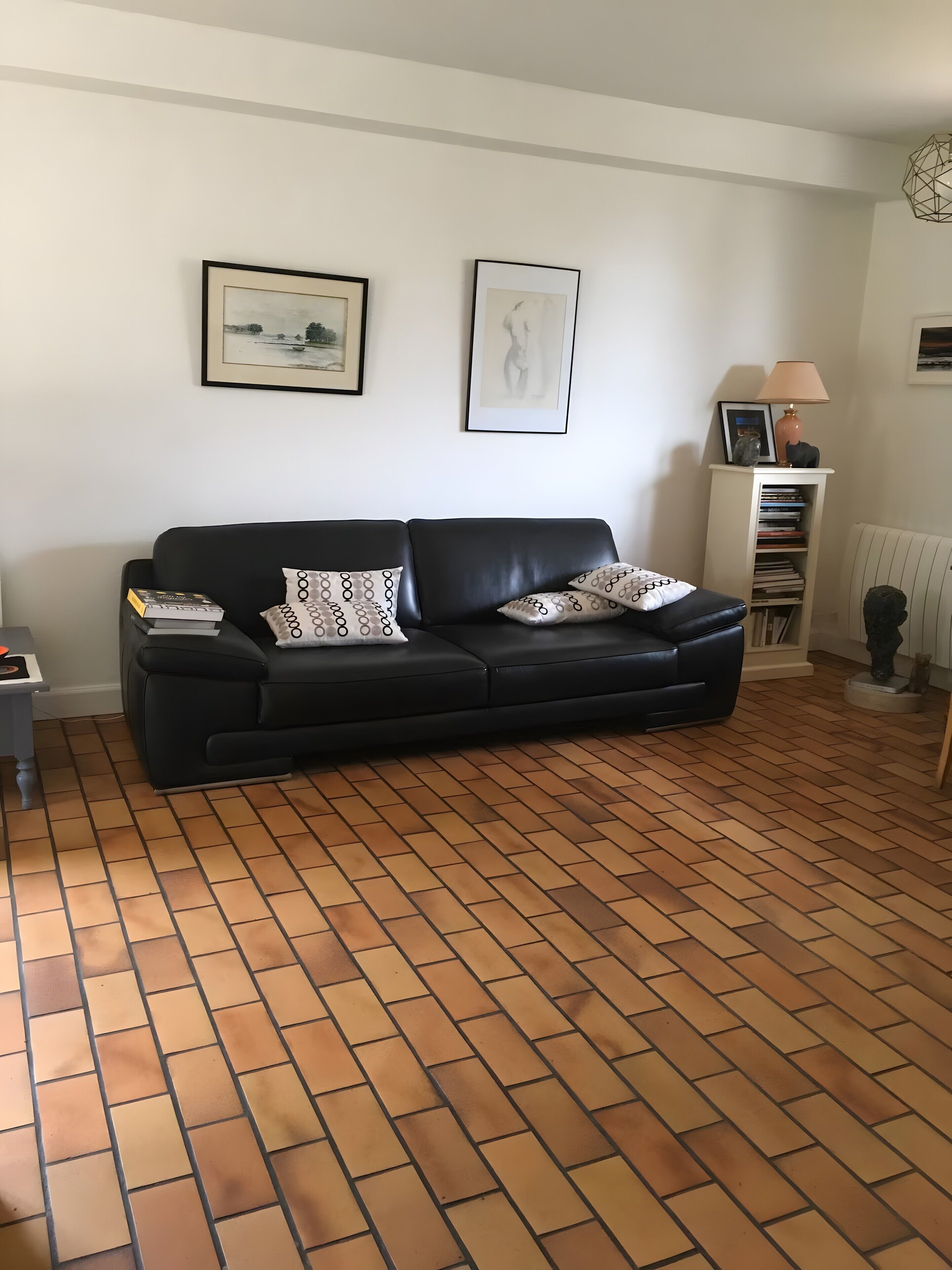 A living room with a black leather sofa, two framed artworks, a lamp, and a radiator.