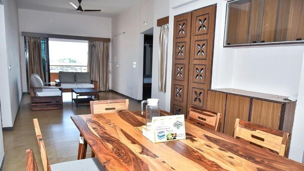 A spacious living area with a wooden dining table, cushioned chairs, a sofa, and a view through a large window.