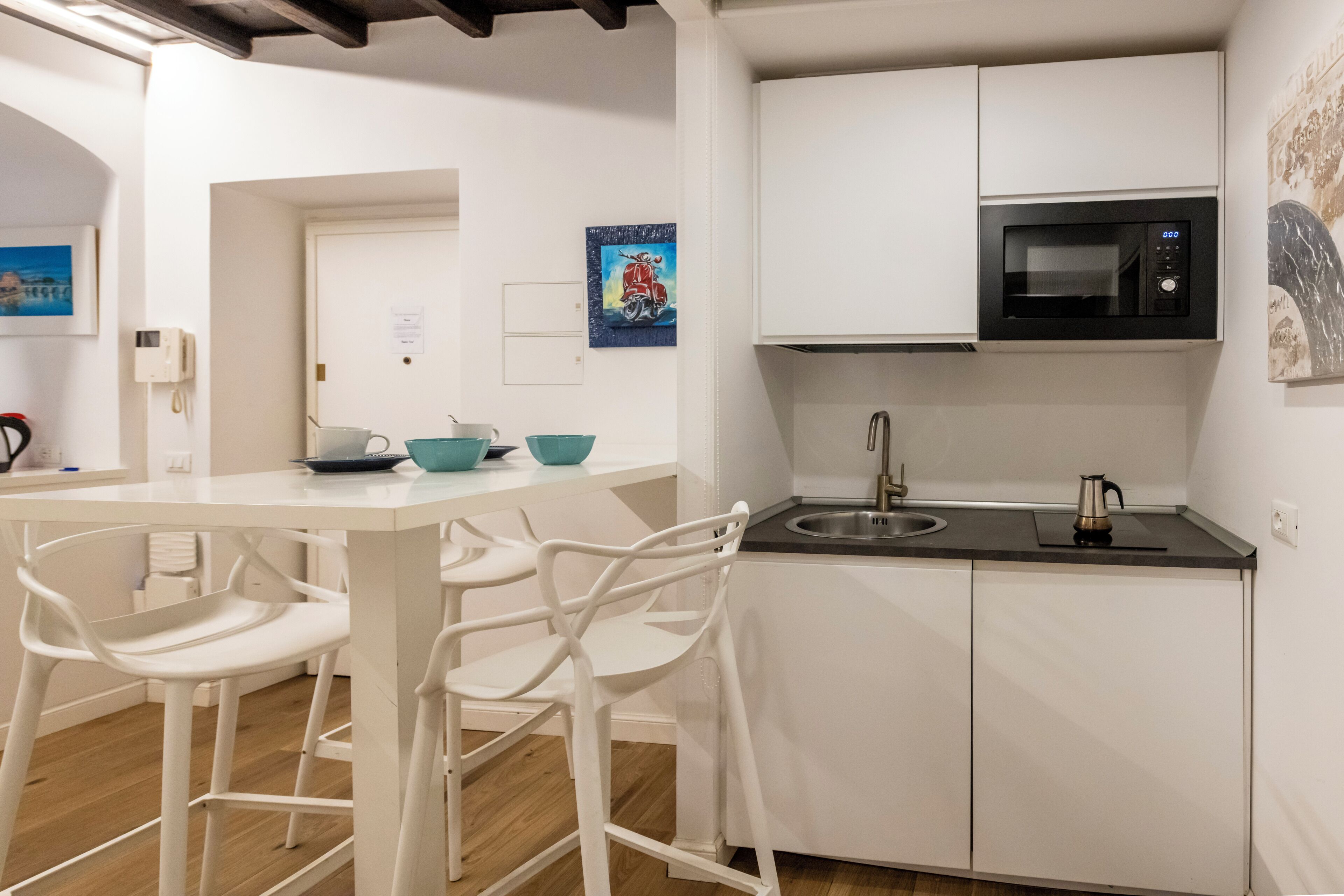A modern kitchen with white cabinetry, a black microwave, and a stainless steel sink. There are white chairs around a table with teal bowls.