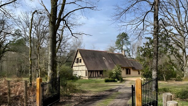 A traditional timber-framed house with a dirt path leading to a gated entrance, surrounded by trees and a grassy area.