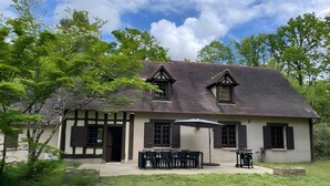 A two-story house with a sloped roof, a front porch with a table and chairs, and a small garden area.