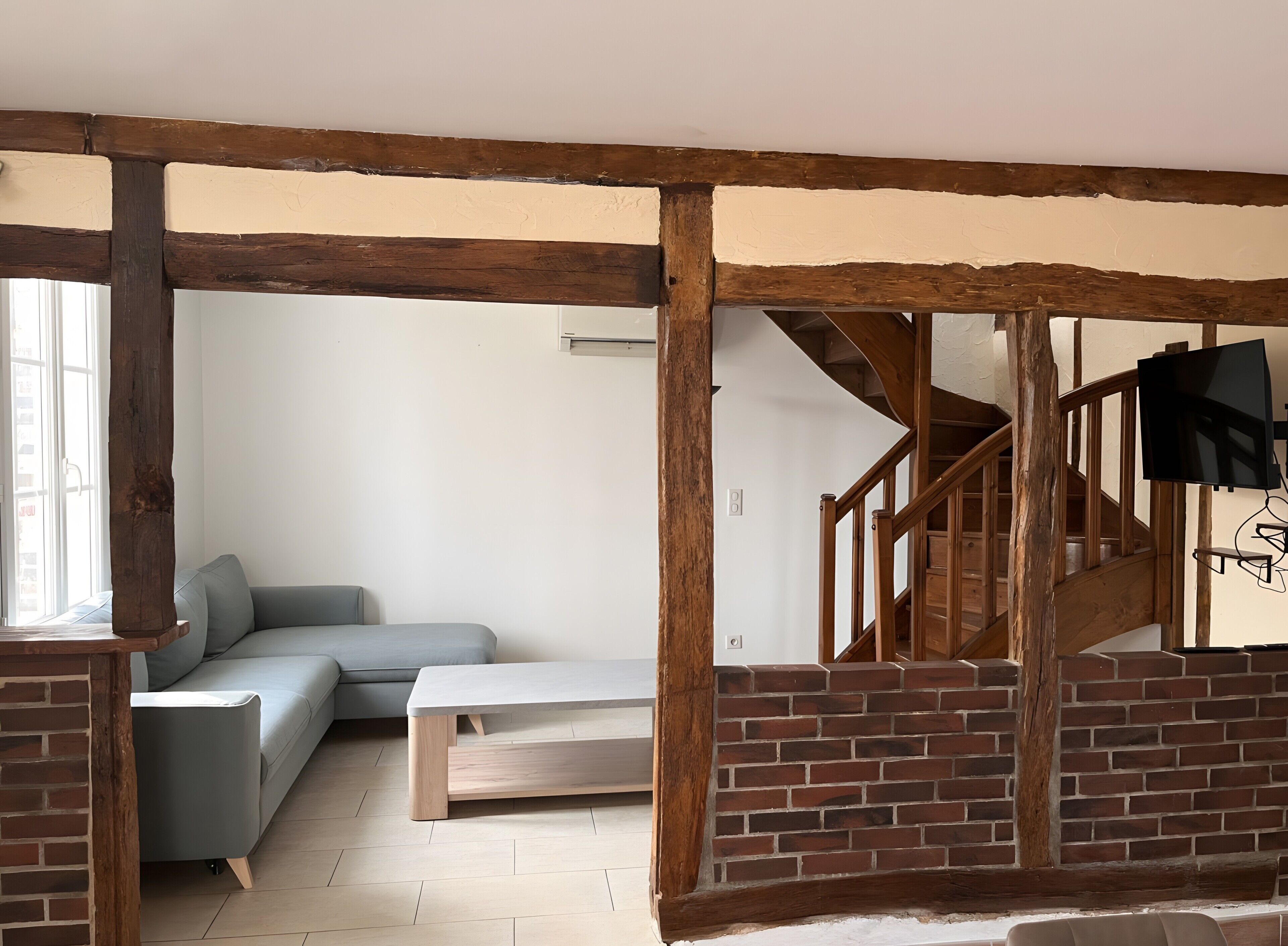 Un salon comprenant un canapé, un banc, des poutres apparentes, un escalier et un téléviseur fixé au mur.