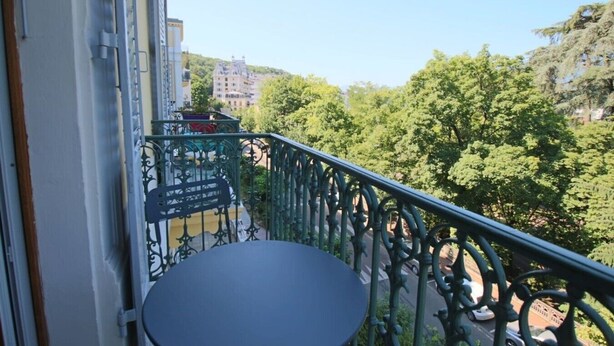 Ein Balkon mit Metallgeländer, einem Tisch und Blick auf einen grünen Park sowie Gebäude.