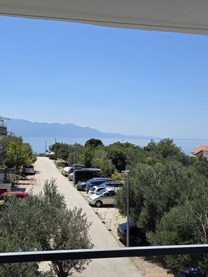 Eine Küstenstraße mit parkenden Autos, Bäumen und freiem Blick auf das Meer und die Berge in der Ferne.